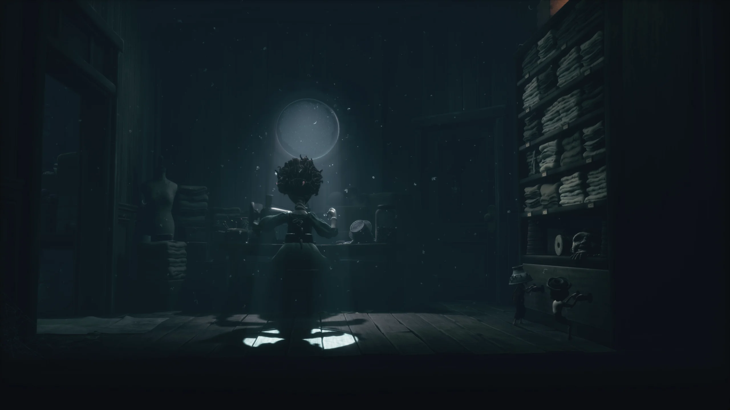 Capture d'écran du contenu additionnel de Little Nightmares III : The Backstage. Une couturière est à l'oeuvre, de dos, dans une pièce très sombre où seul une légère lumière provient d'une petite fenêtre, mettant en valeur la femme en plein travail.